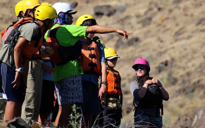 A group of people wearing helmets and life jackets stand on land. One of them points outward.